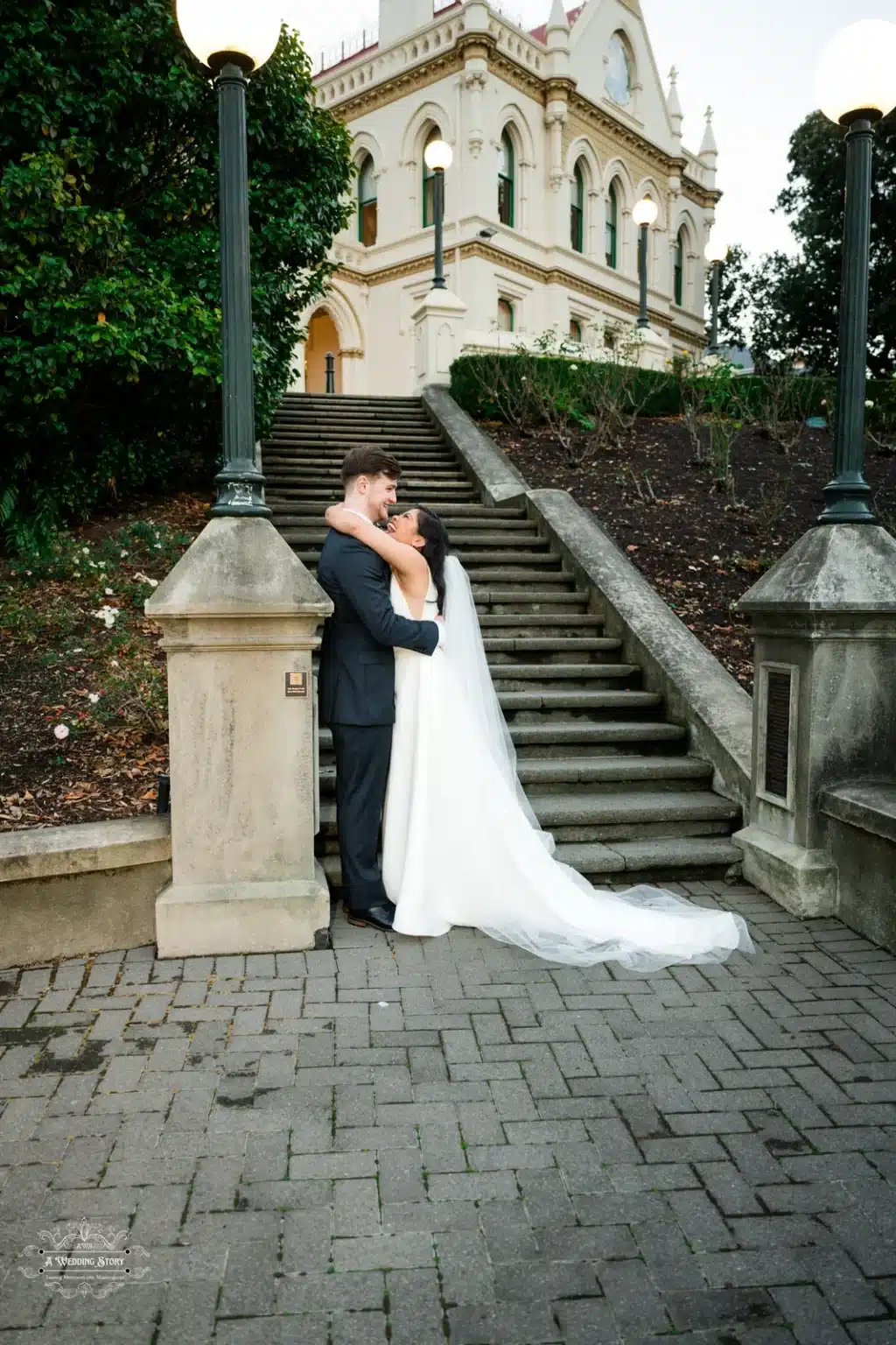 Bride and groom embracing on historic stone stairs, surrounded by elegant lamp posts and lush greenery.