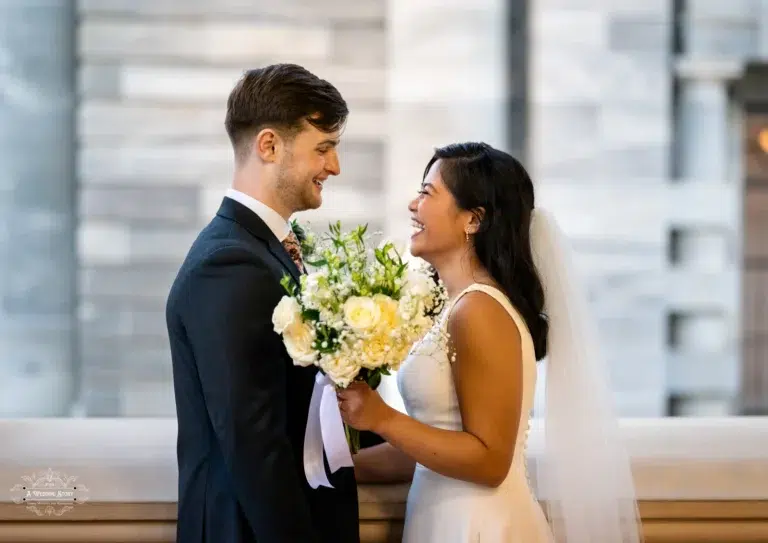 A happy bride and groom smiling at each other while holding a bouquet of white and yellow roses in Wellington, New Zealand.