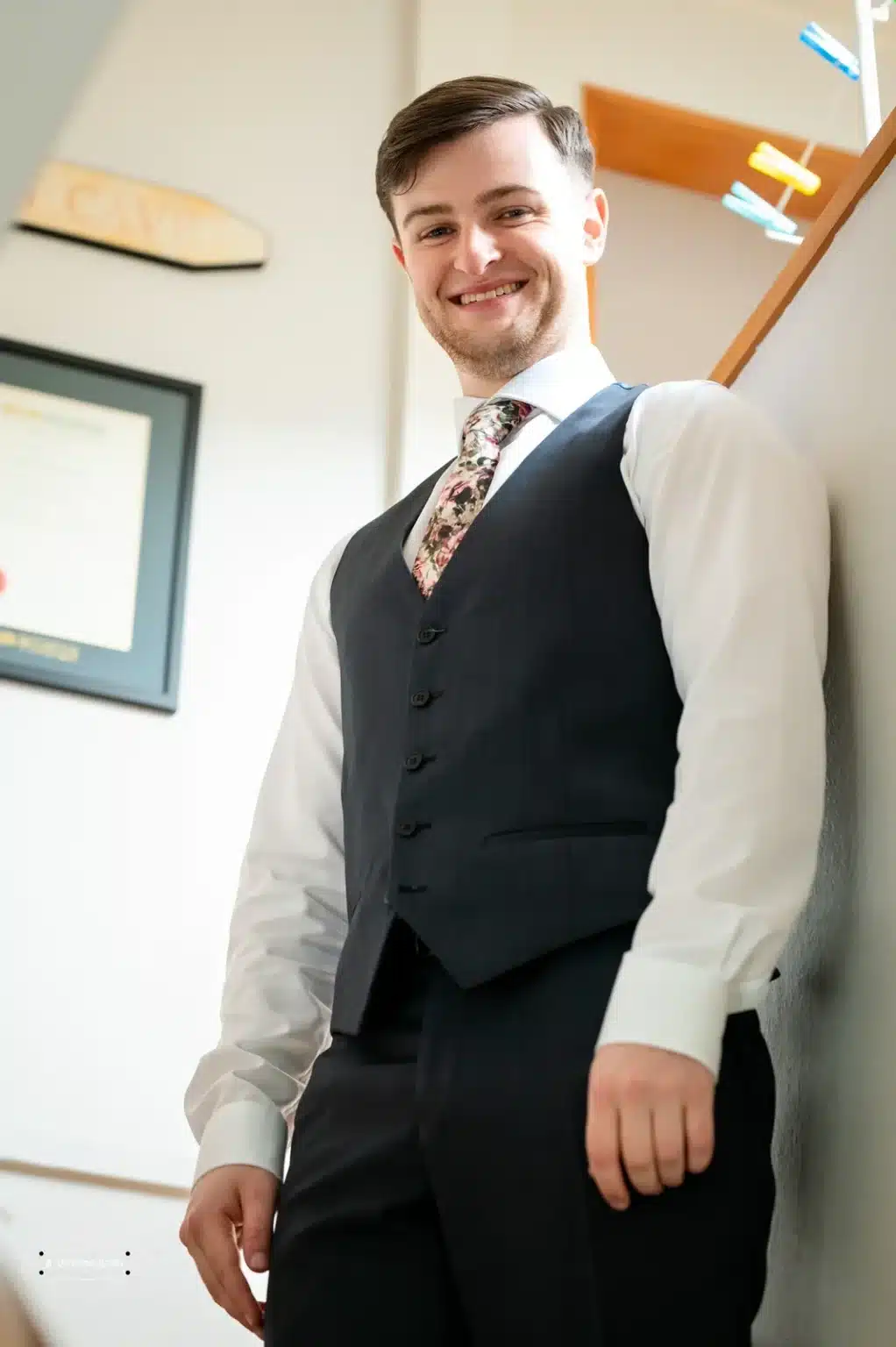 A cheerful groom dressed in a tailored three-piece suit with a floral tie, standing confidently and smiling brightly on his wedding day in Wellington, New Zealand.