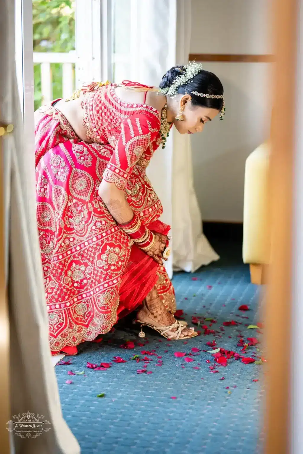 Gujarati bride in a red and gold embroidered lehenga adjusting her anklet, with scattered rose petals on the floor in a serene indoor setting.