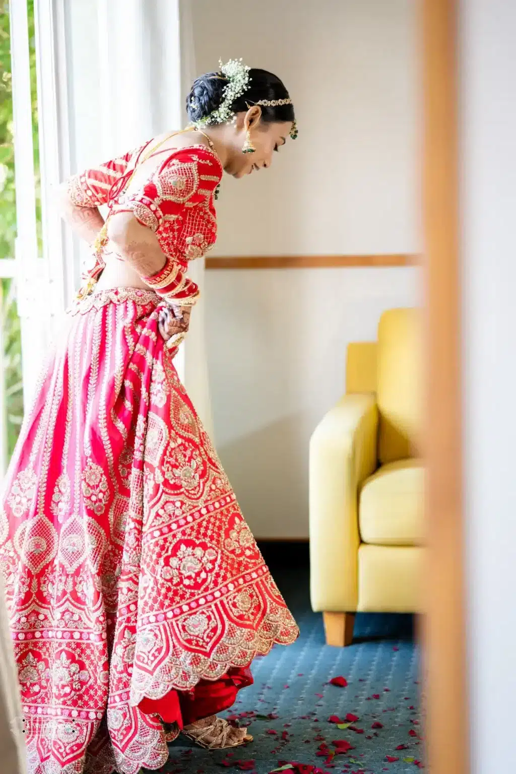 Gujarati bride in a red lehenga adjusting her outfit, with golden embroidery and scattered rose petals on the floor, captured in a softly lit room.