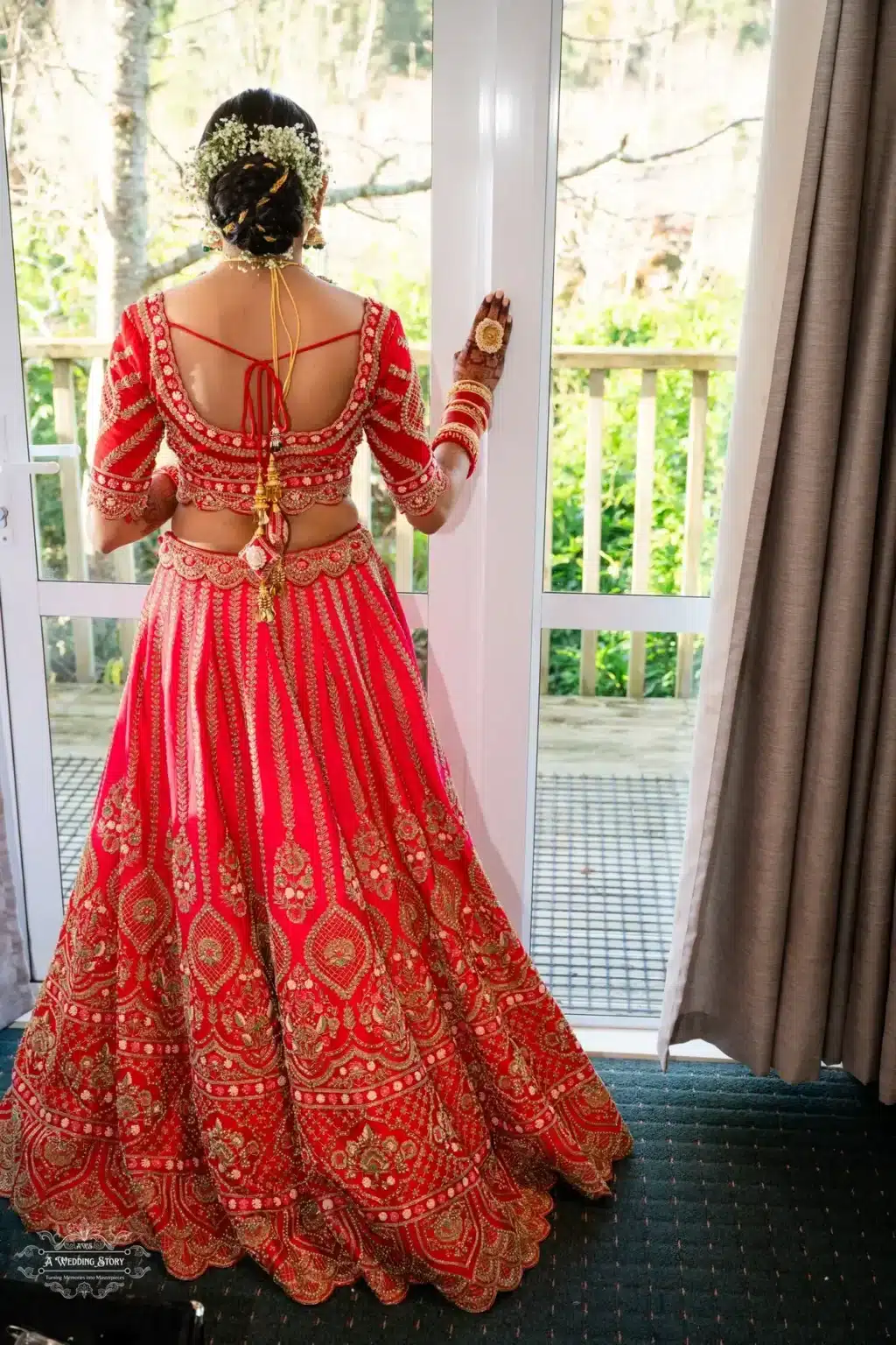 Gujarati bride in a red lehenga, showcasing intricate embroidery and a bridal hairstyle adorned with floral accessories, captured near a glass door.