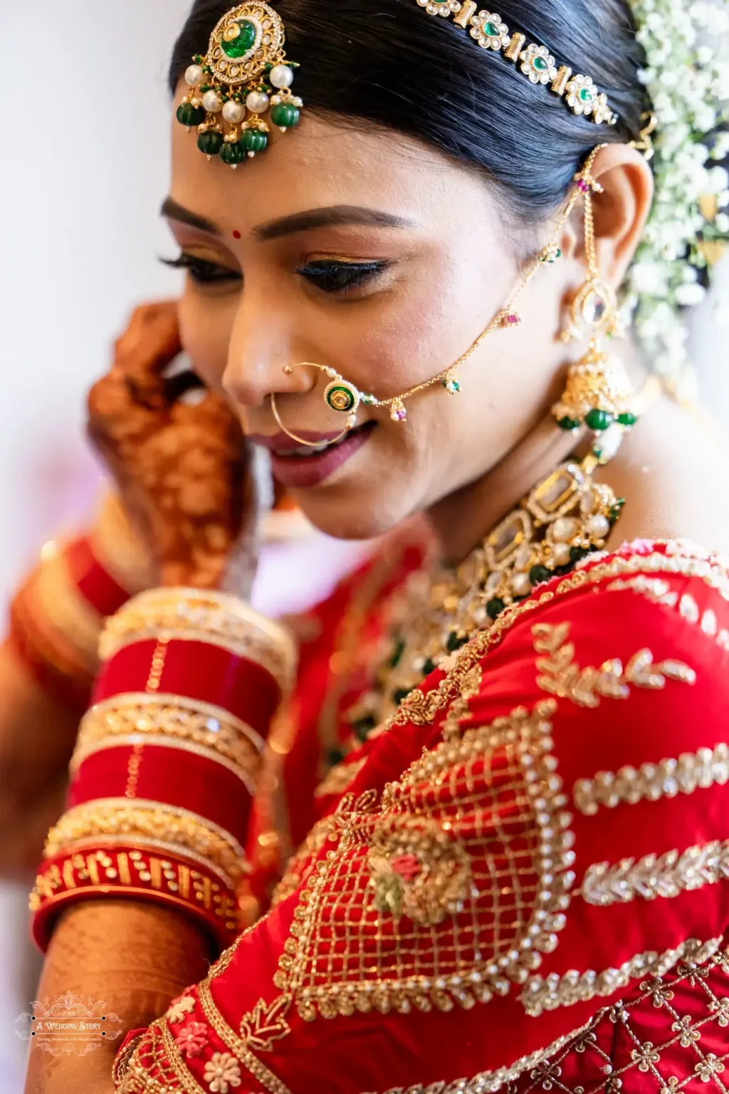 Gujarati bride in a traditional red blouse adorned with intricate embroidery, adjusting her earrings. The image highlights her green-stoned maang tikka, bridal jewelry, and henna-adorned hands.