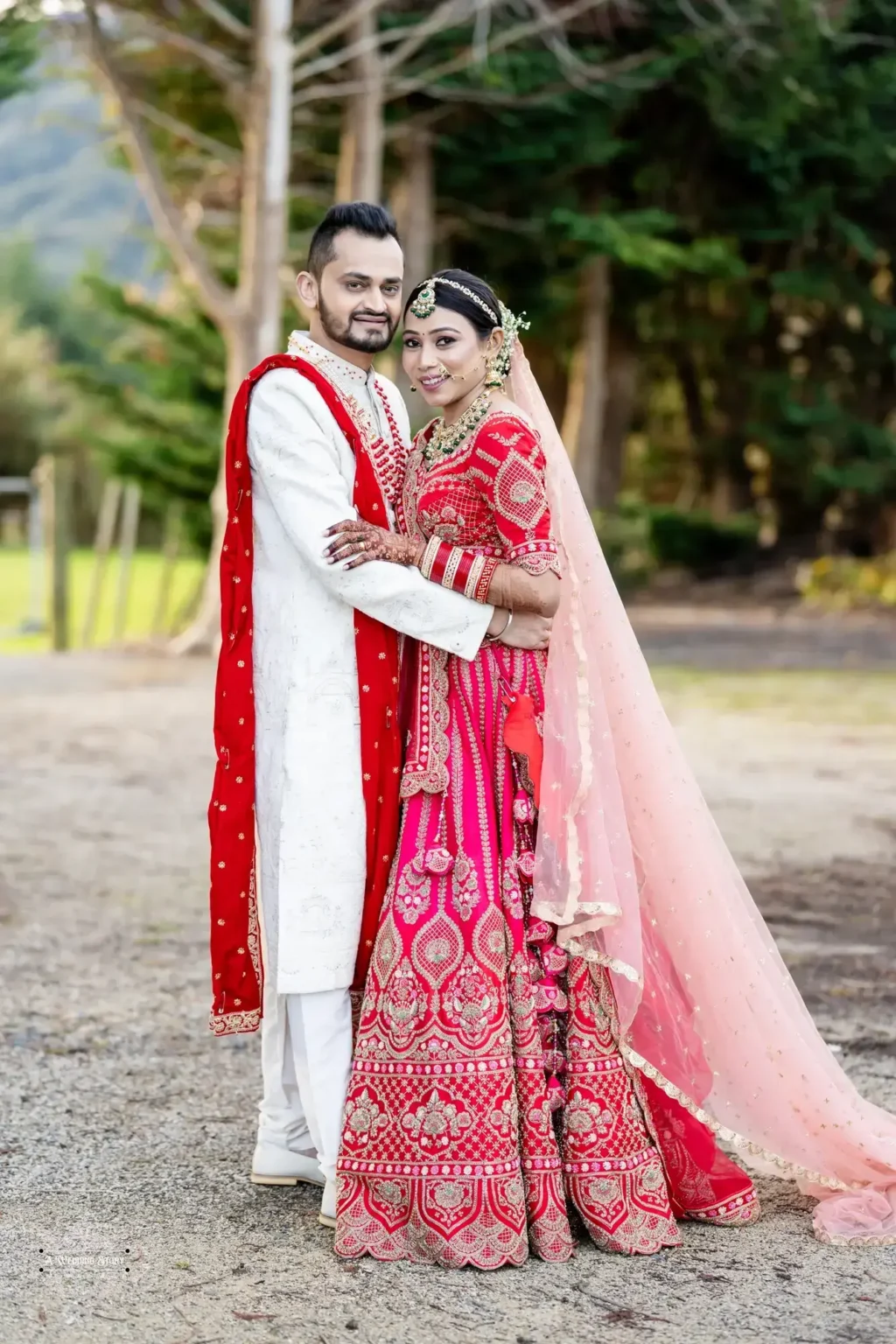 Smiling Gujarati bride in a red lehenga and groom in a white sherwani with red accents embracing during their outdoor wedding photoshoot in Wellington, New Zealand