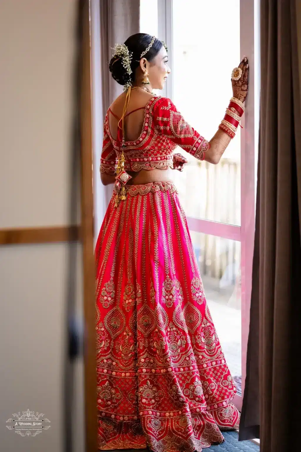 Gujarati bride standing near a glass door, wearing a traditional red lehenga with golden embroidery and a floral-adorned bridal hairstyle, showcasing elegance and intricate details.