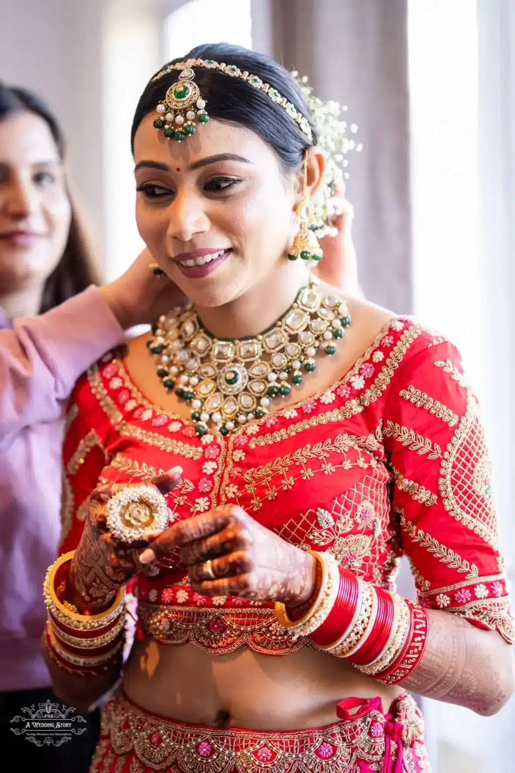 A Gujarati bride dressed in a red lehenga with golden embroidery, wearing traditional jewelry, as her stylist adjusts her bridal necklace.