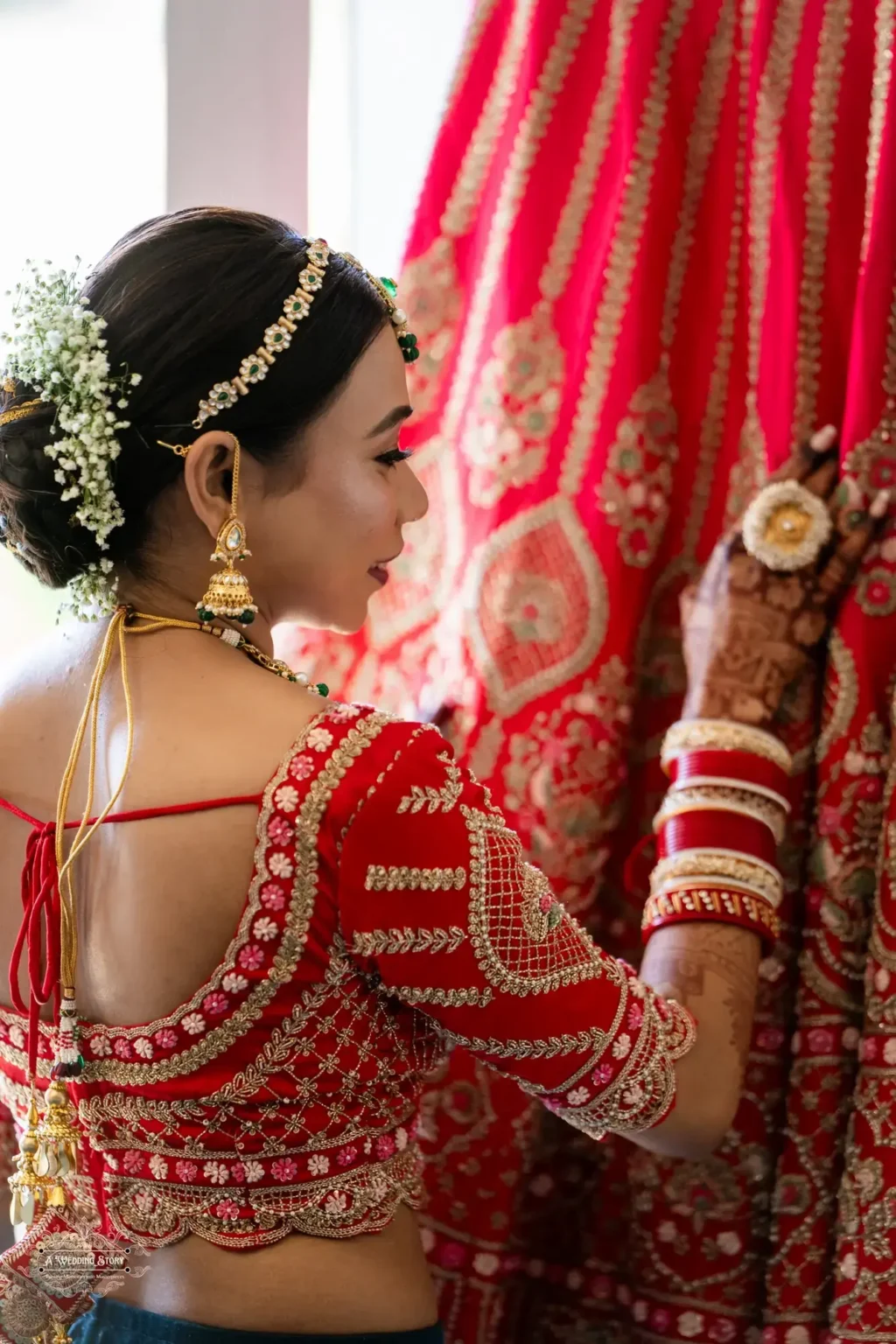 Gujarati bride's side profile showcasing intricate golden embroidery on her red bridal blouse, adorned with jewelry and floral hair accessories, while gently touching her lehenga.