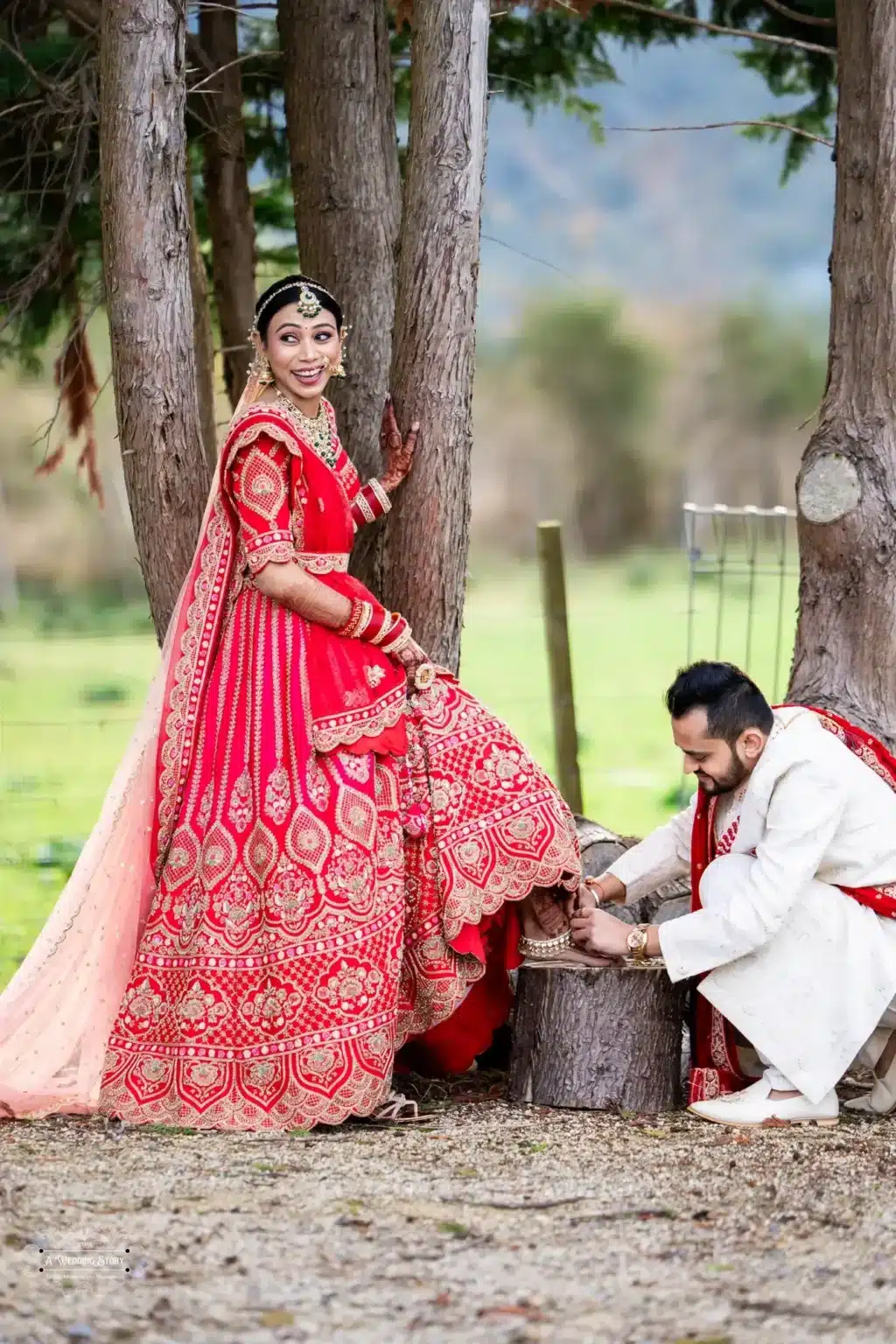 Smiling Gujarati bride in a vibrant red lehenga and groom in a white sherwani adjusting her anklet during their outdoor wedding in Wellington, New Zealand