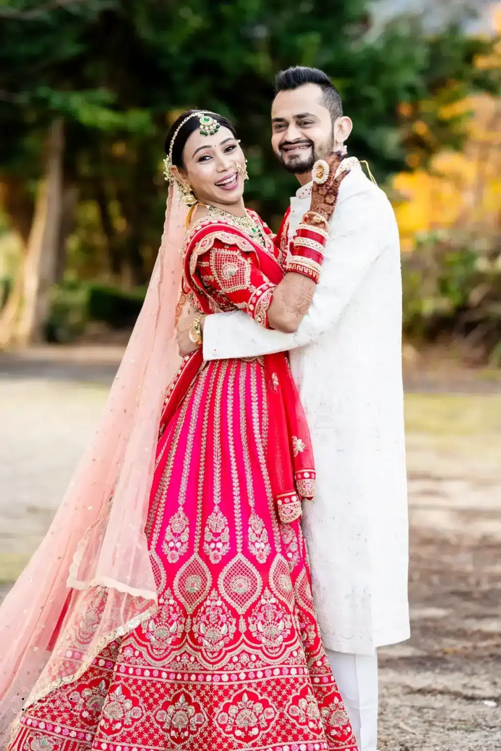 Indian bride in a vibrant red lehenga and groom in a white sherwani sharing a joyful embrace during their Gujarati wedding photoshoot in Wellington, New Zealand