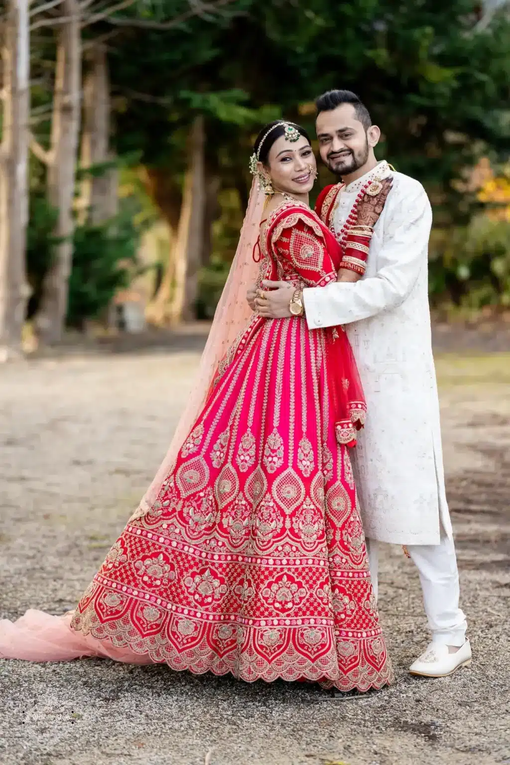 Smiling Gujarati bride in a red lehenga and groom in a white sherwani sharing a loving embrace during their traditional wedding photoshoot in Wellington, New Zealand