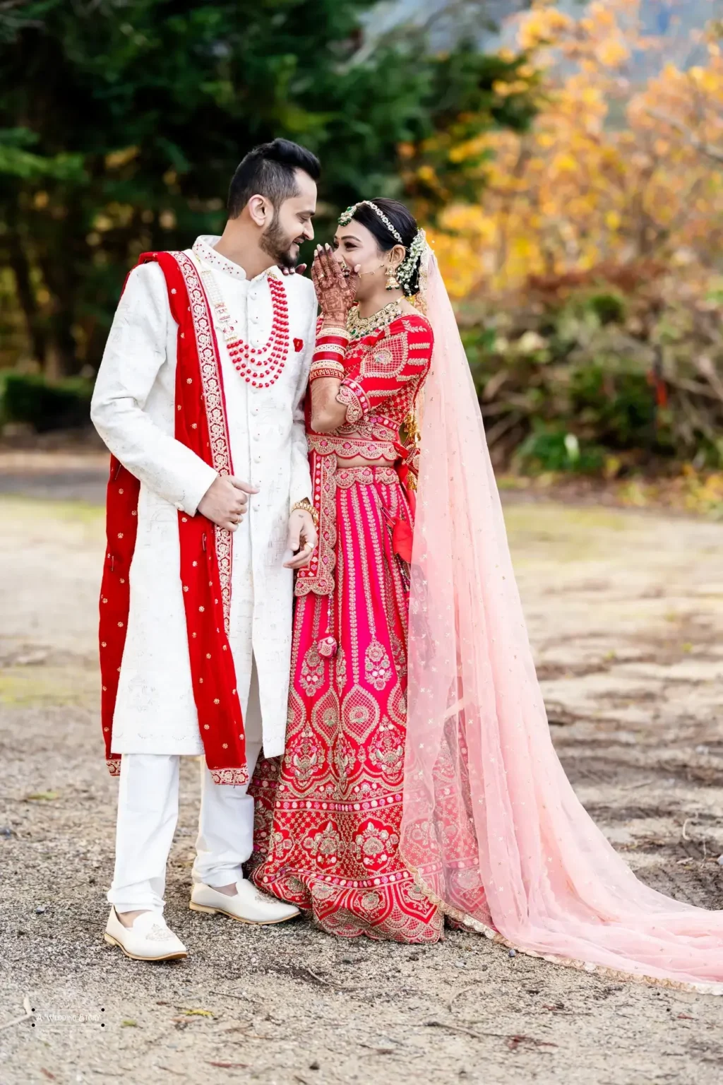 Smiling Gujarati bride in a red lehenga sharing a playful moment with the groom in a white sherwani during their wedding photoshoot in Wellington, New Zealand