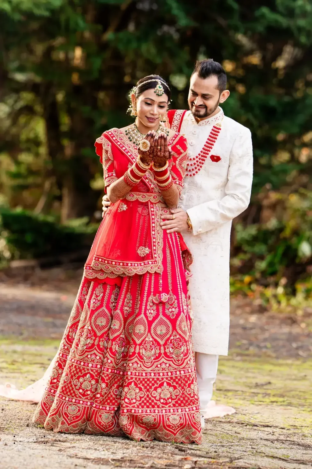 Gujarati bride in a red lehenga and groom in a white sherwani sharing a tender moment while admiring her henna during their wedding photoshoot in Wellington, New Zealand