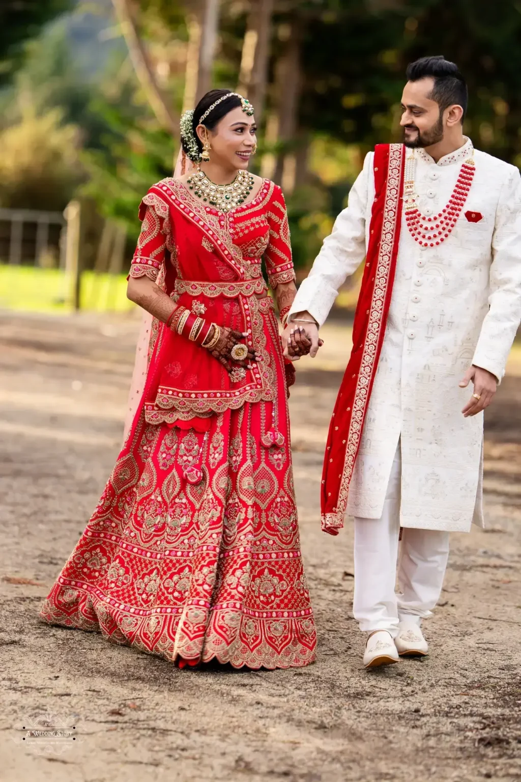 Smiling Gujarati bride in a red lehenga and groom in a white sherwani holding hands during their wedding photoshoot in Wellington, New Zealand.