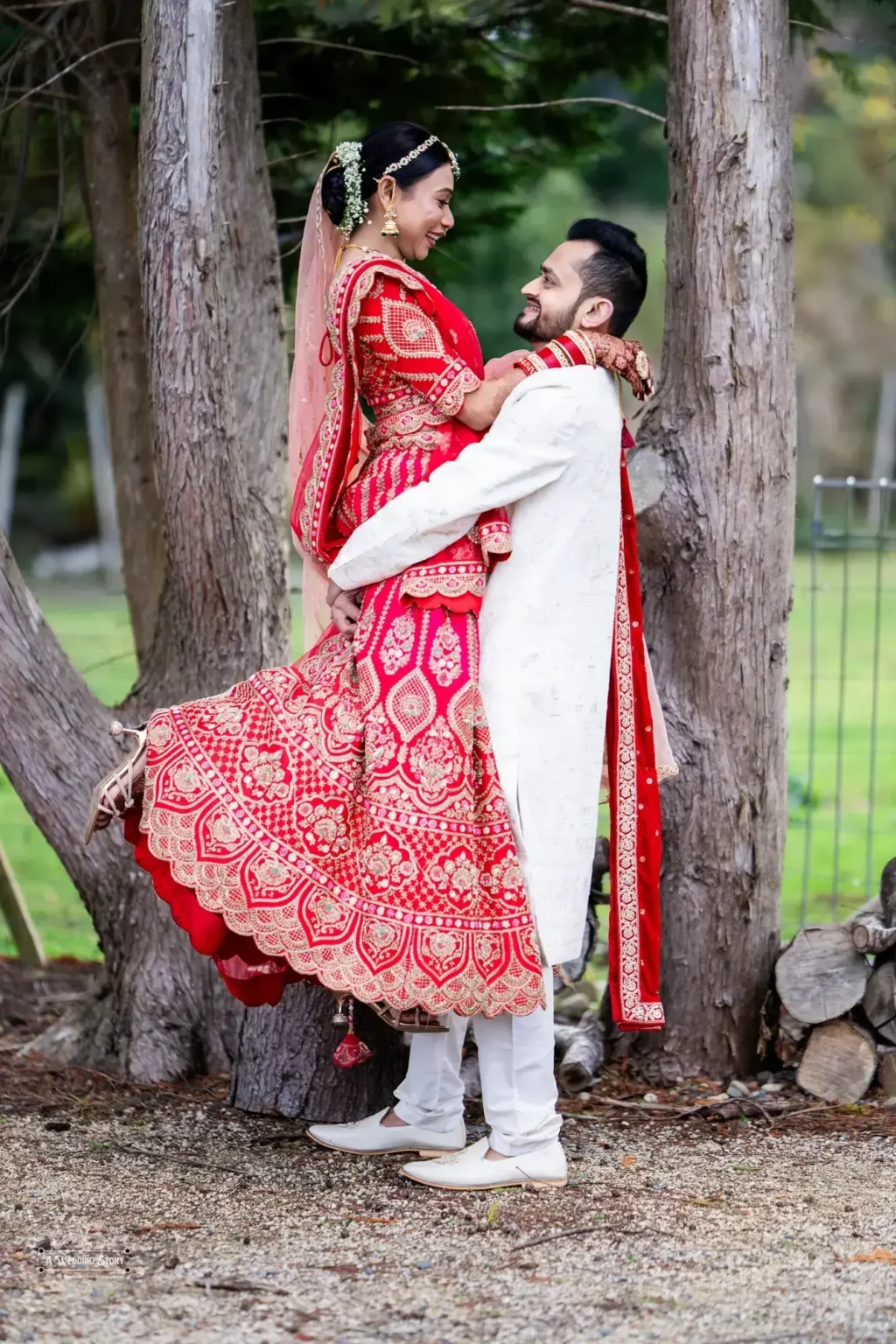 Gujarati groom in a white sherwani lifting his bride in a vibrant red lehenga during their wedding photoshoot in Wellington, New Zealand.