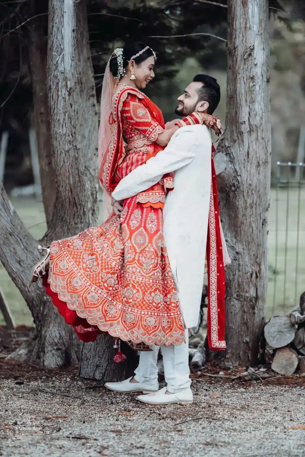 Gujarati groom in a white sherwani lifting his bride in a vibrant red lehenga during a candid moment of their wedding photoshoot in Wellington, New Zealand.