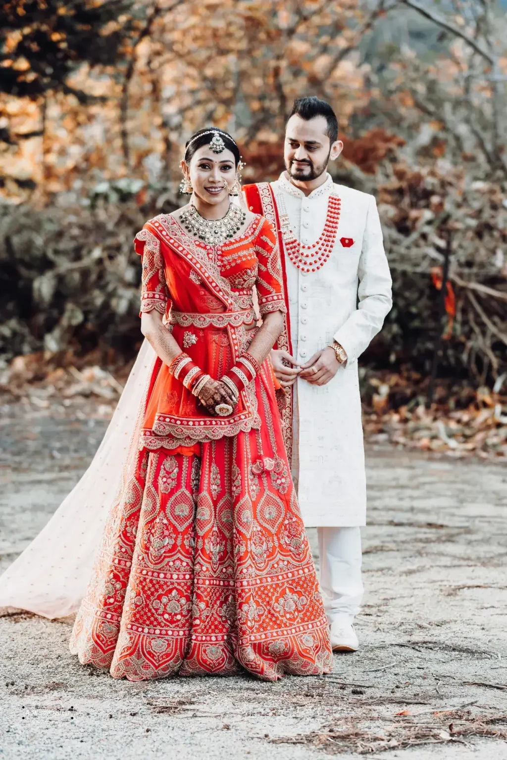 Gujarati bride in a red lehenga and groom in a white sherwani posing during their wedding photoshoot in Wellington, New Zealand, against a fall foliage backdrop