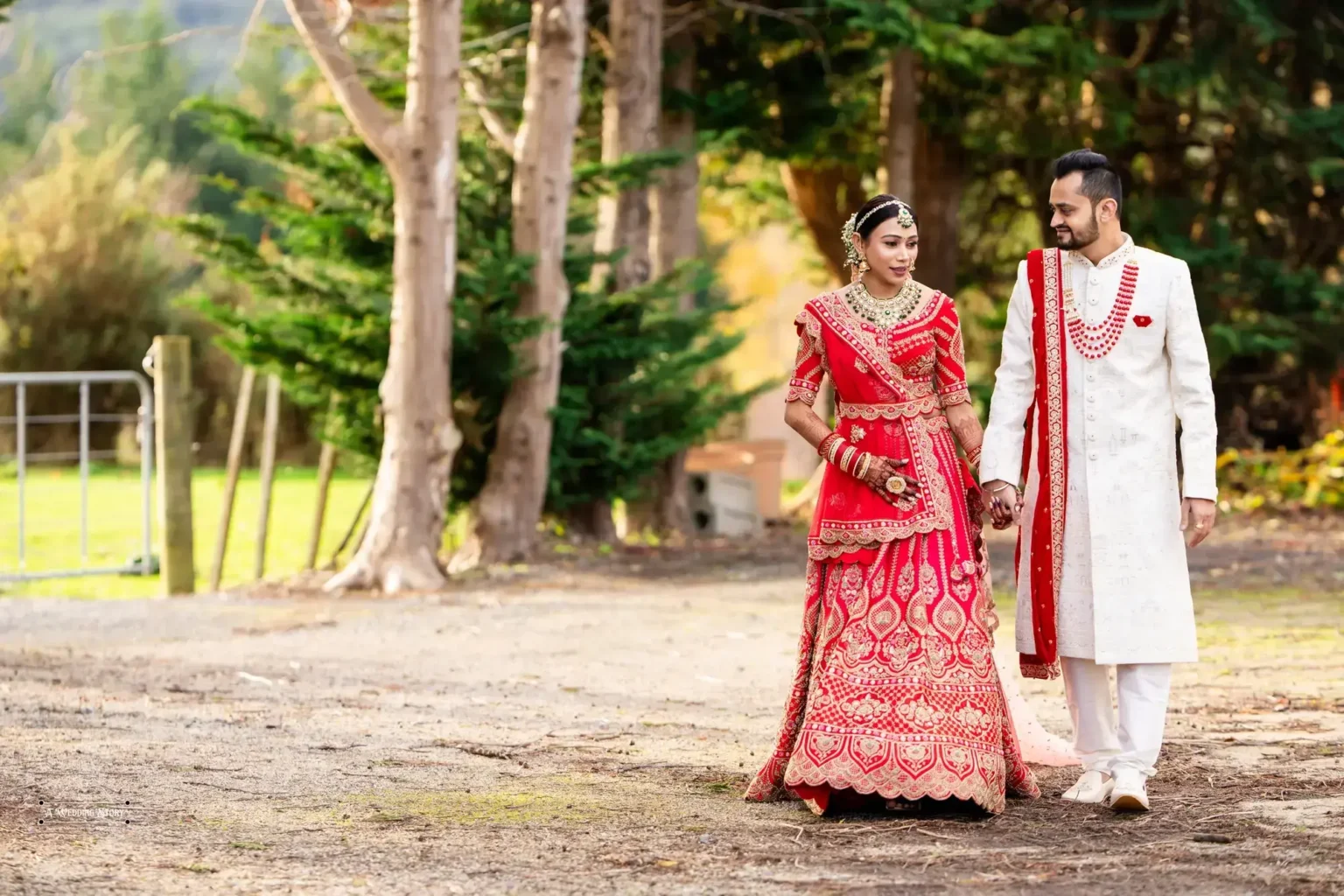 Gujarati bride in a vibrant red lehenga and groom in a white sherwani walking hand-in-hand during their wedding photoshoot in Wellington, New Zealand