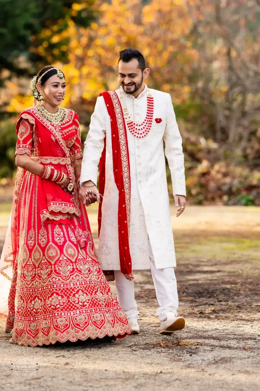 Happy Gujarati bride in a red lehenga and groom in a white sherwani walking hand-in-hand during their wedding photoshoot in Wellington, New Zealand.