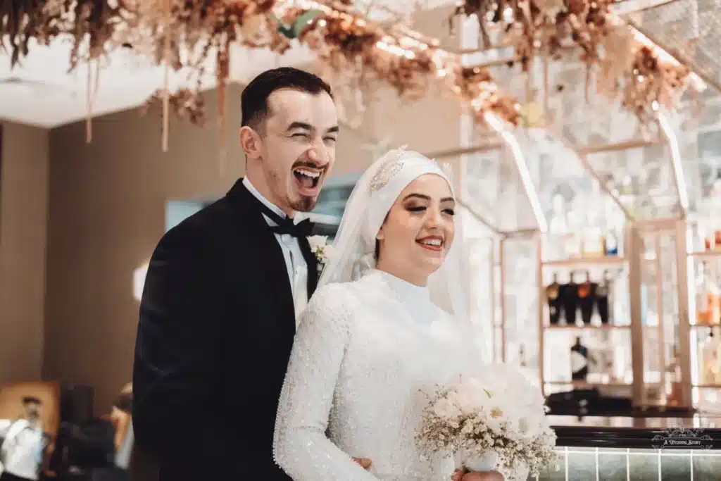 Afghan bride in a white hijab and groom in a tuxedo laughing together during their wedding celebration in Wellington.