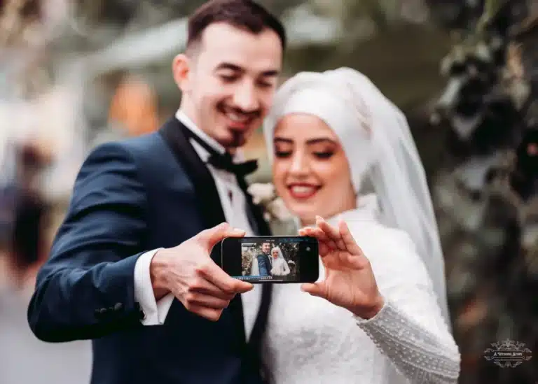 Afghan bride in a traditional white hijab and groom in a tuxedo taking a selfie on their wedding day, smiling and celebrating outdoors in Wellington.