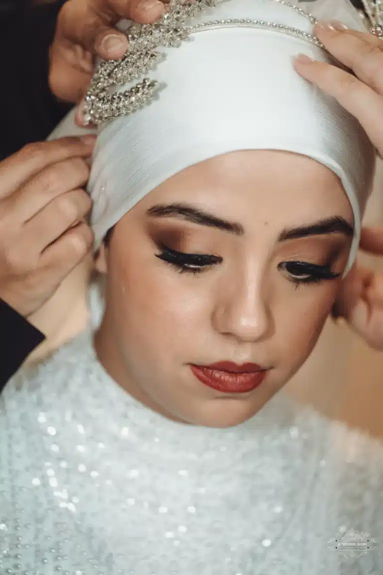 Afghan bride in a white hijab with an intricate headpiece, focused as hands adjust her wedding attire in Wellington.