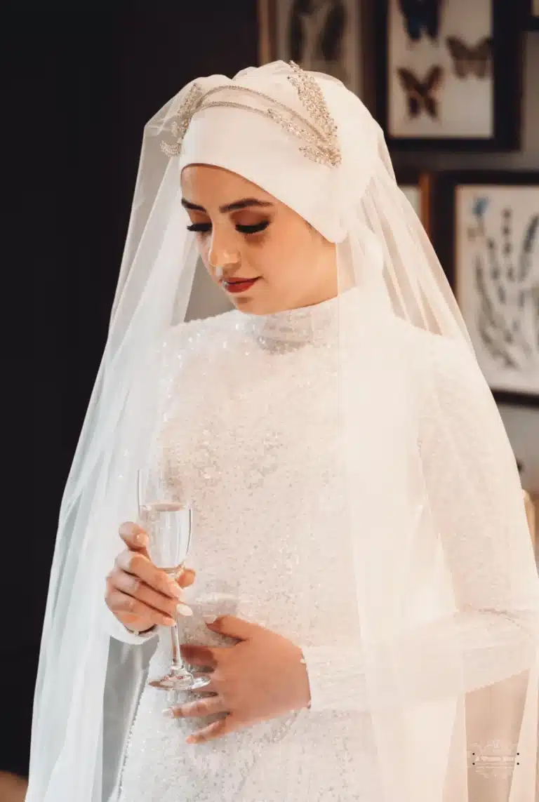 Afghan bride in a white beaded gown and headpiece, holding a glass and looking down thoughtfully during her wedding in Wellington.