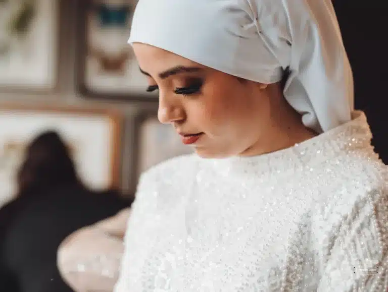 Afghan bride in a white hijab and beaded gown, looking down thoughtfully as she prepares for her wedding day in Wellington.