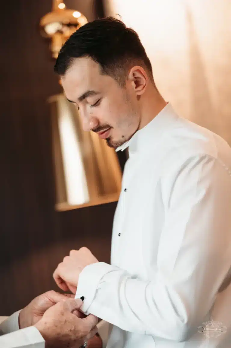 Afghan groom in a white dress shirt, receiving assistance with his cufflink, captured in a thoughtful moment before his wedding in Wellington.