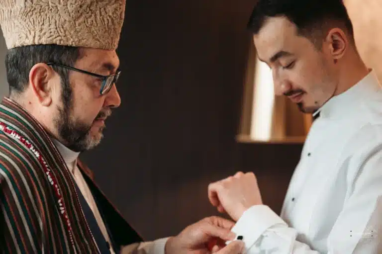 Afghan groom in a white dress shirt receiving assistance with his cufflink from an elder family member wearing traditional attire before his wedding in Wellington.