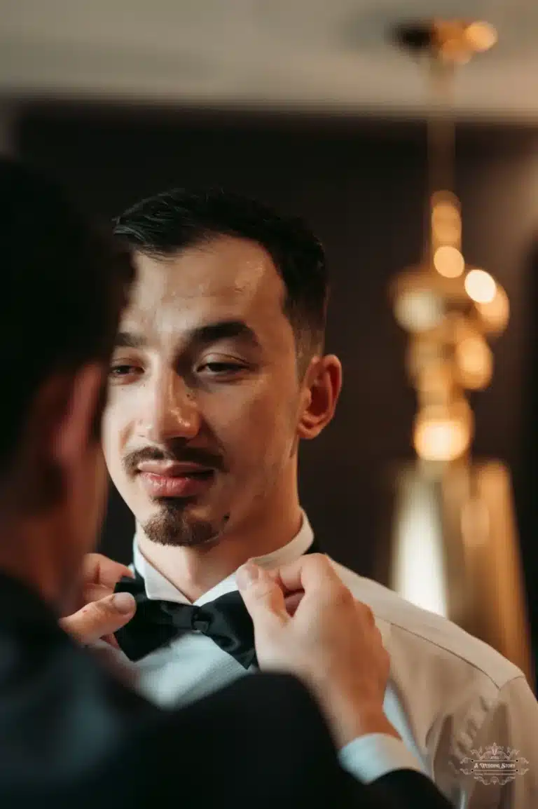 Afghan groom in a tuxedo, receiving assistance with his bow tie, preparing for his wedding in Wellington.