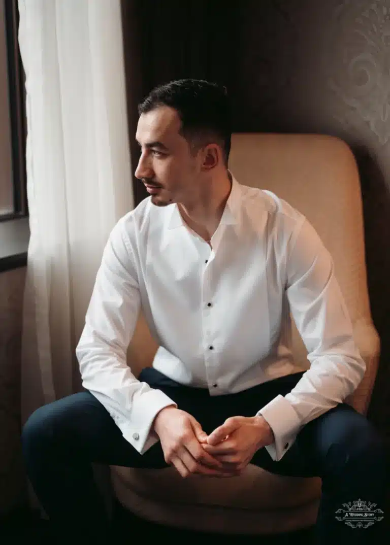 Afghan groom in a white dress shirt, seated by the window, looking thoughtfully to the side on his wedding day in Wellington.