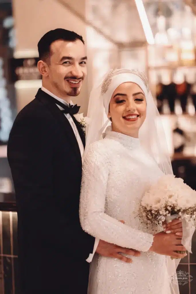 Afghan bride in a white hijab holding a bouquet, smiling beside her groom in a tuxedo during their wedding celebration in Wellington.