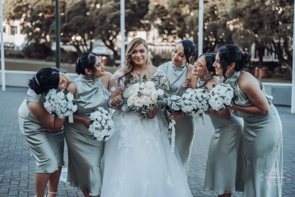 Bride surrounded by her bridesmaids holding white bouquets, sharing a joyful and lighthearted moment during a wedding photoshoot in Wellington, New Zealand.