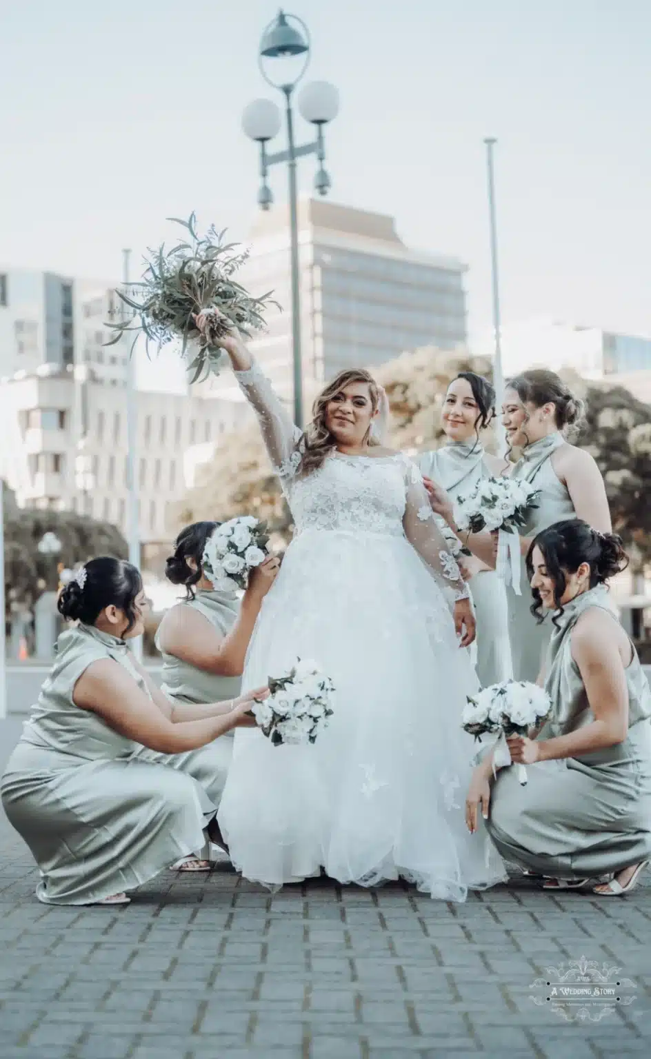 Bride raising her bouquet triumphantly while surrounded by her bridesmaids holding white flowers during a wedding photoshoot in Wellington, New Zealand.