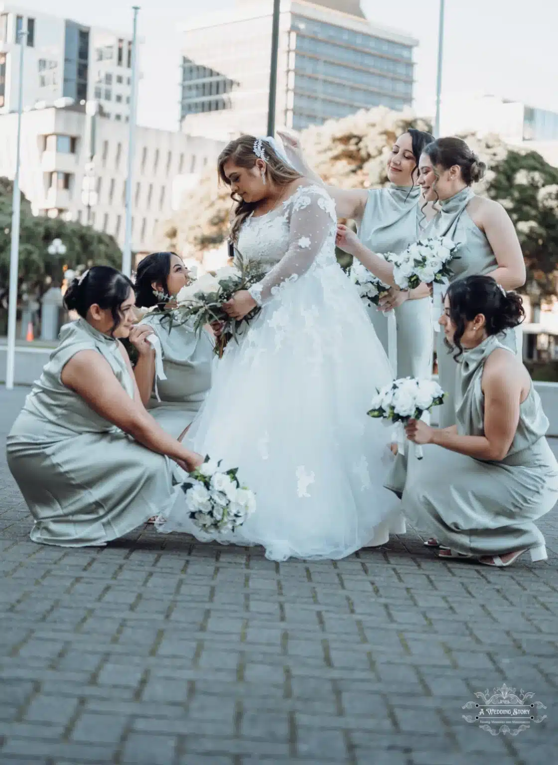 Bride standing gracefully as her bridesmaids adjust her dress and veil while holding white bouquets during a wedding photoshoot in Wellington, New Zealand.