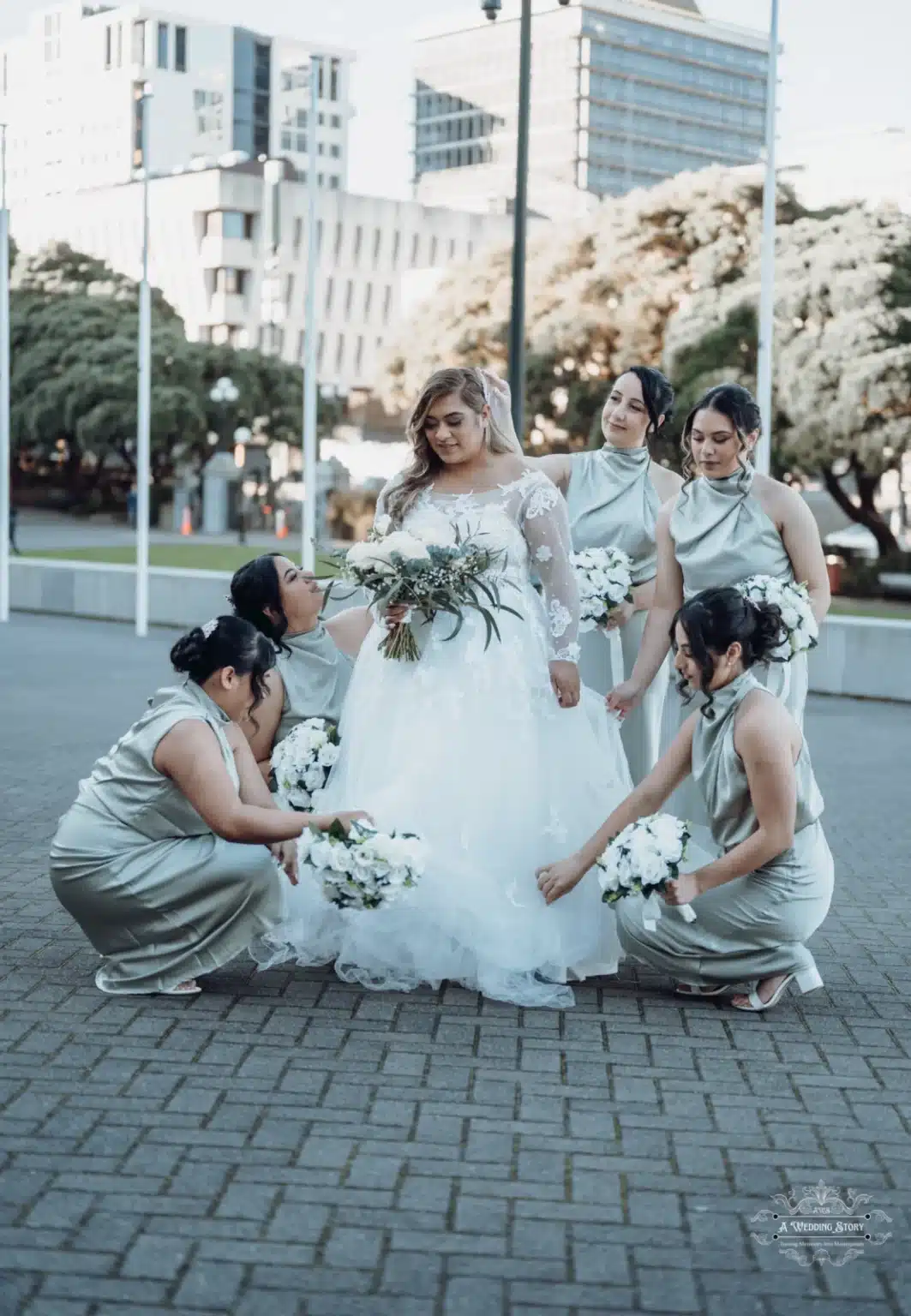 Bride standing gracefully as her bridesmaids adjust her dress and hold bouquets during a wedding photoshoot in Wellington, New Zealand.