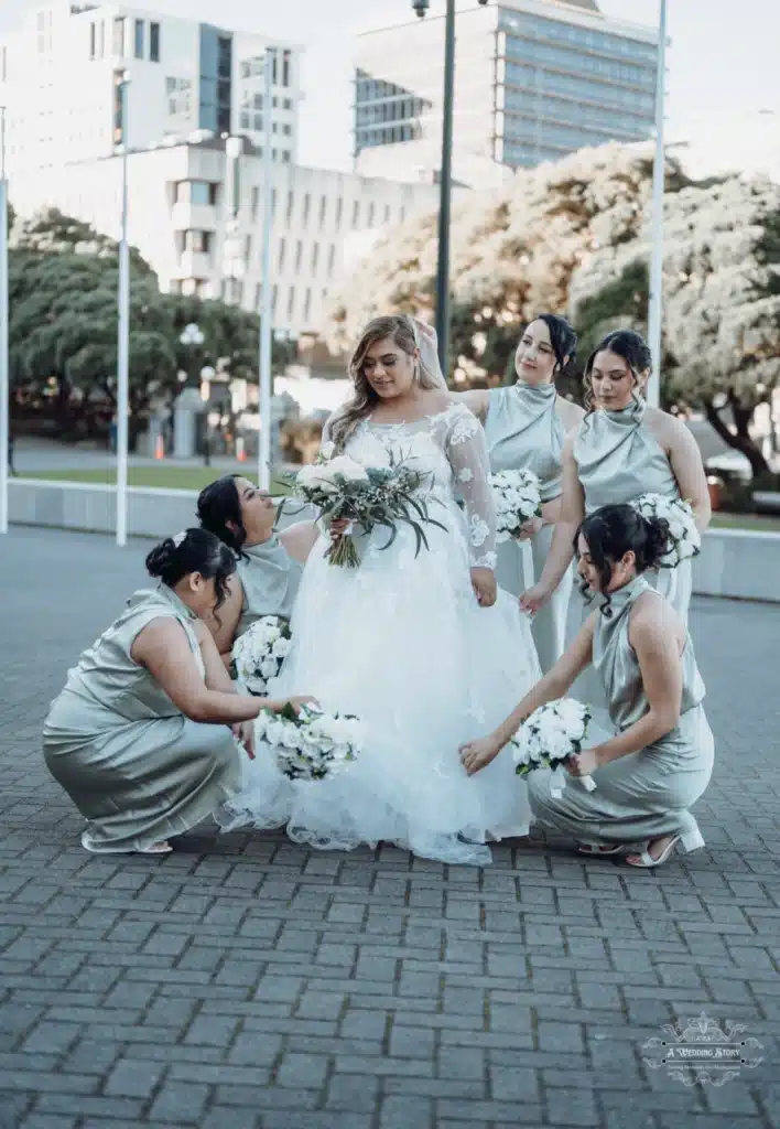 Bride standing gracefully as her bridesmaids adjust her dress and hold bouquets during a wedding photoshoot in Wellington, New Zealand.