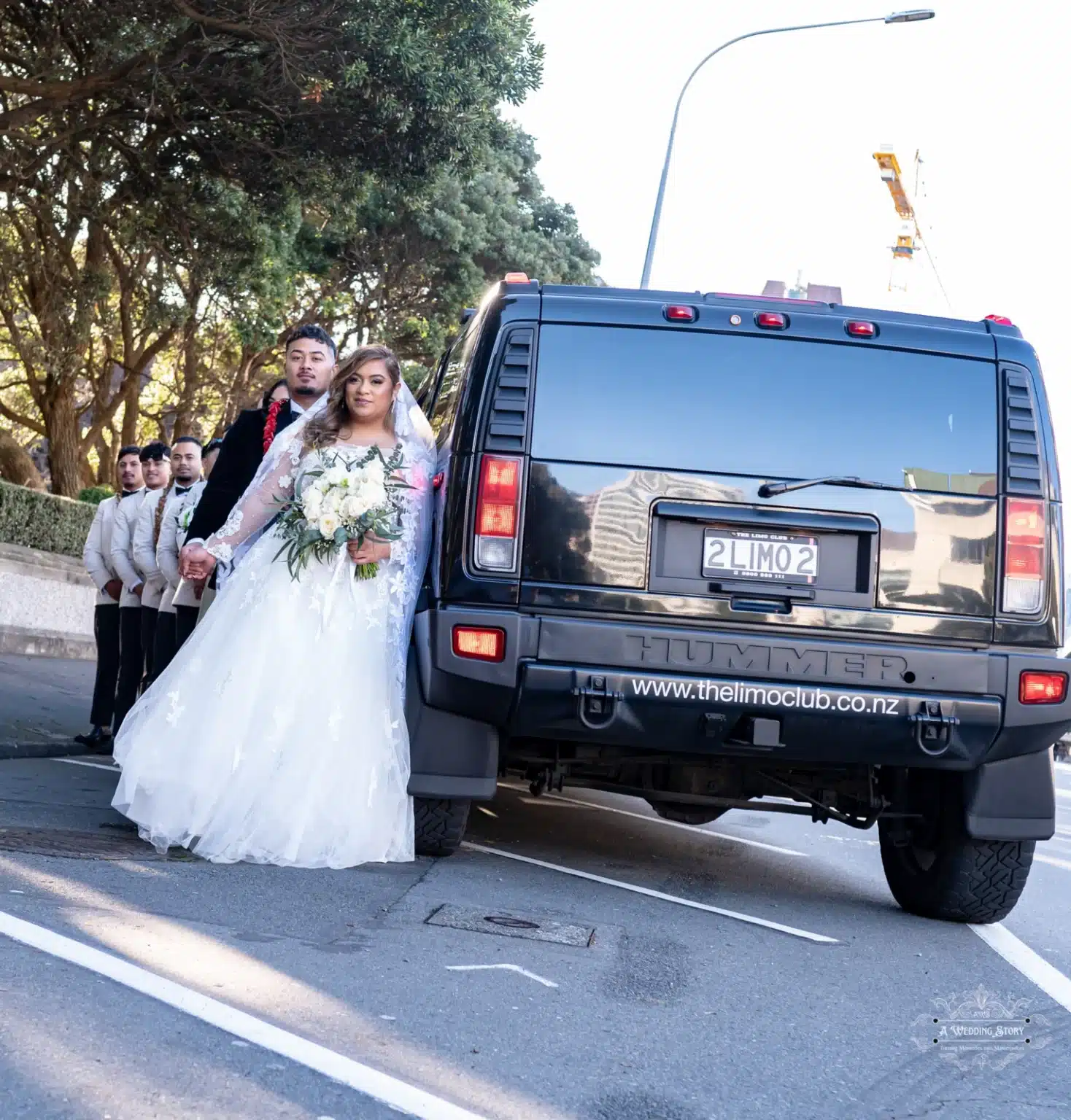 The bride and groom stand hand in hand beside a black Hummer limousine, surrounded by groomsmen in cream suits, under the trees on a sunny Wellington street.