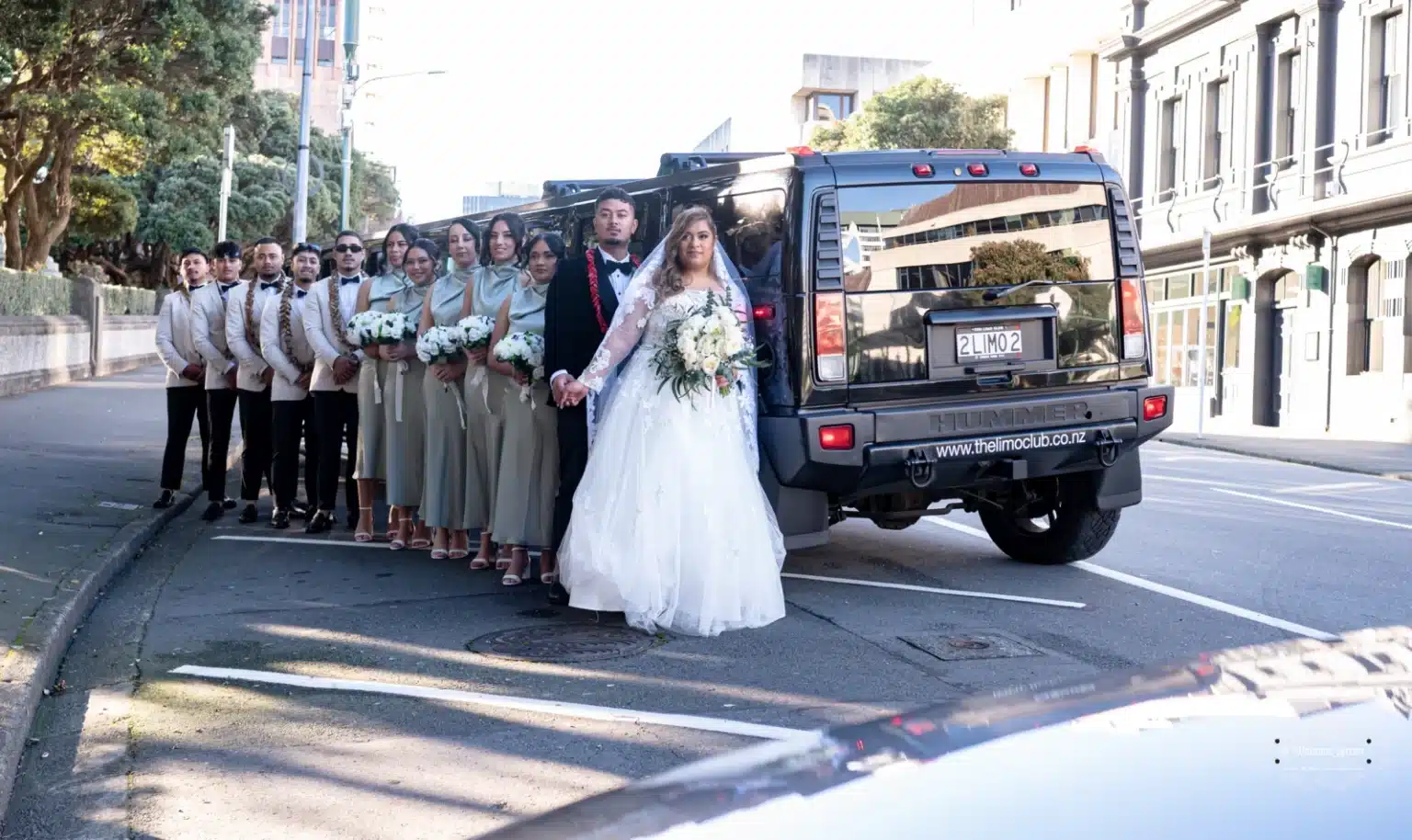 The bride and groom posing with their bridal party alongside a black luxury Hummer limousine during a wedding photoshoot in Wellington, New Zealand.