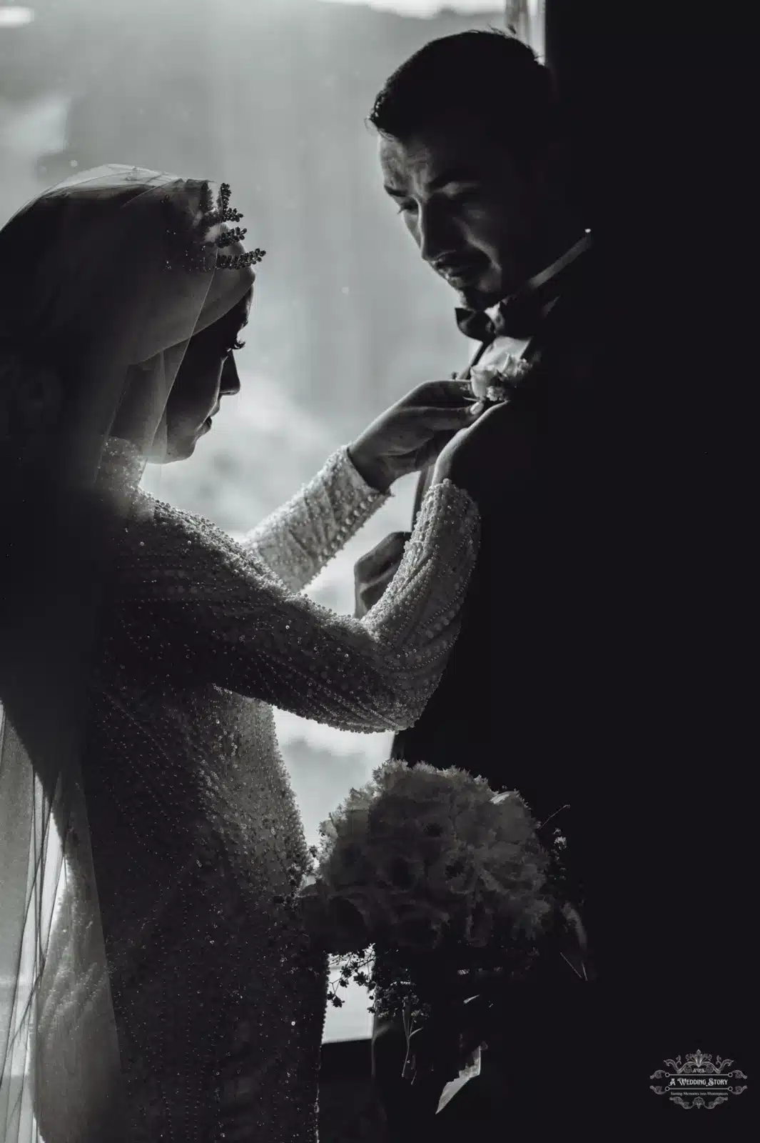 A black-and-white portrait of a bride and groom in a tender moment, with the bride adjusting the groom's boutonniere, captured in soft lighting by a window.