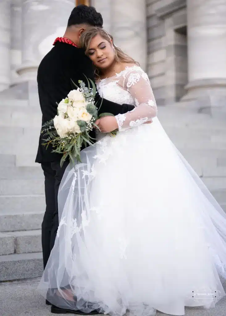 A bride rests her head on the groom’s shoulder, holding a bouquet of white flowers during an intimate wedding photoshoot in Wellington, New Zealand.