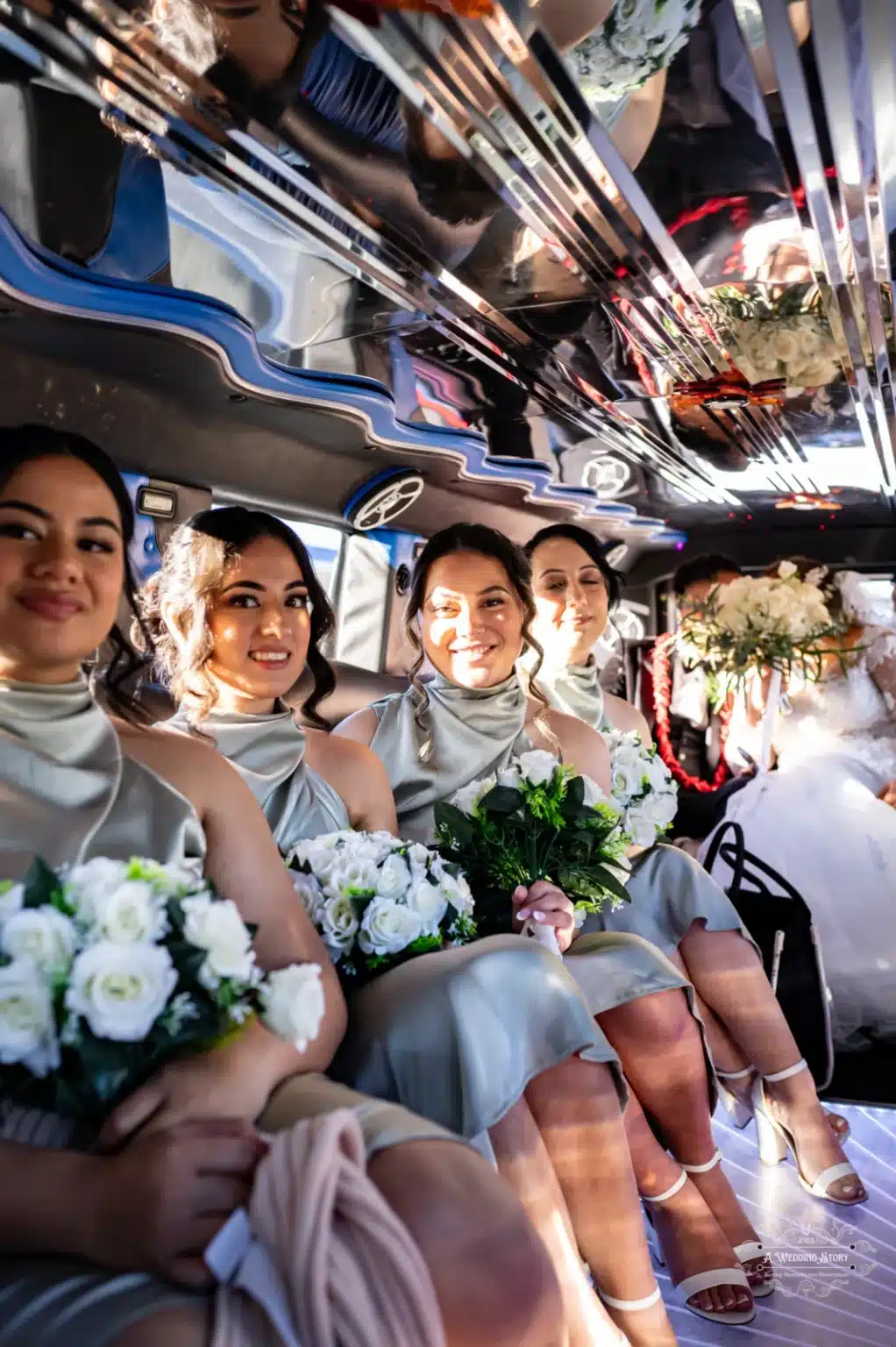 Bridesmaids seated in a decorated limousine, holding white rose bouquets and wearing light satin dresses, with the bride and groom visible in the background celebrating.