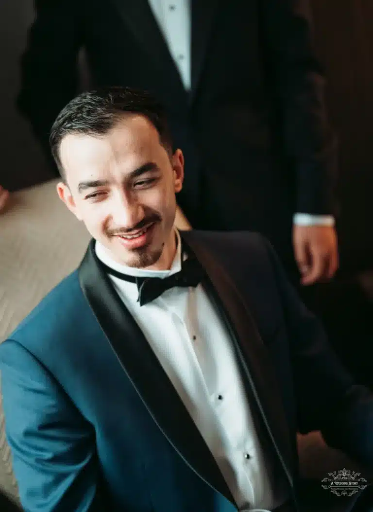 A groom seated and smiling with his best man standing behind, both in formal attire before the wedding ceremony.