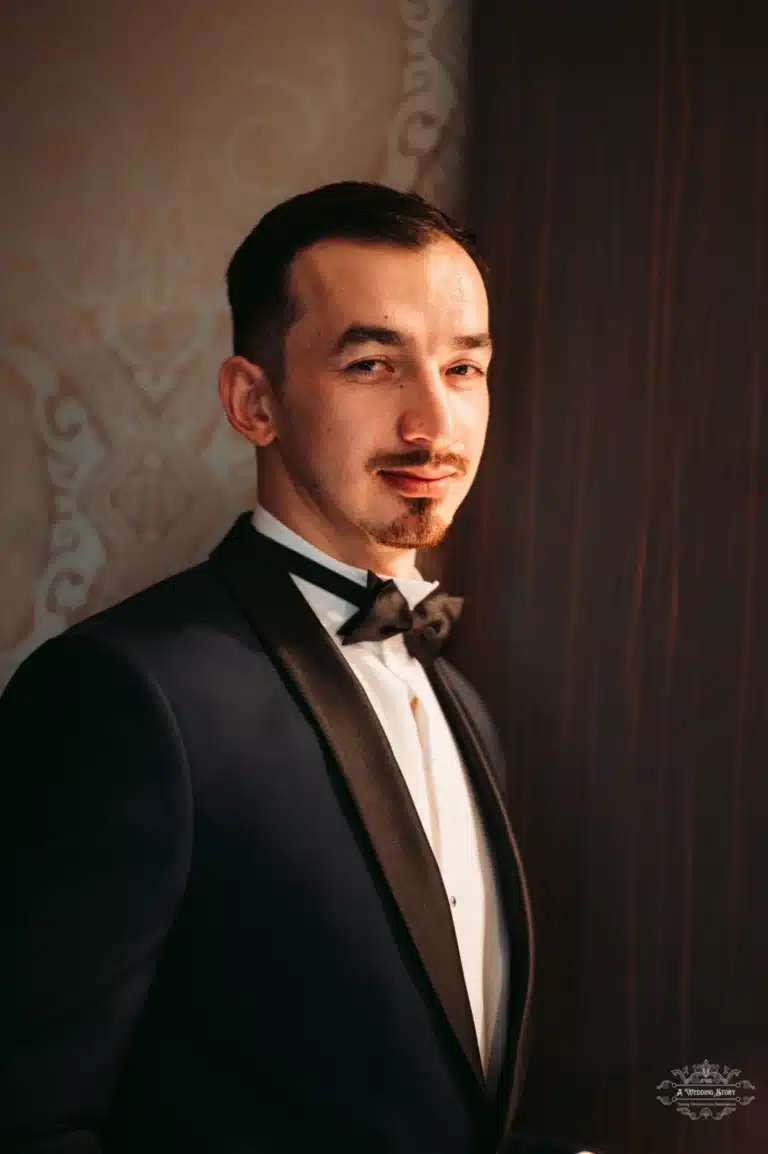 The groom, dressed in a dark tuxedo and bow tie, standing confidently with a subtle smile, highlighted by soft, warm lighting.