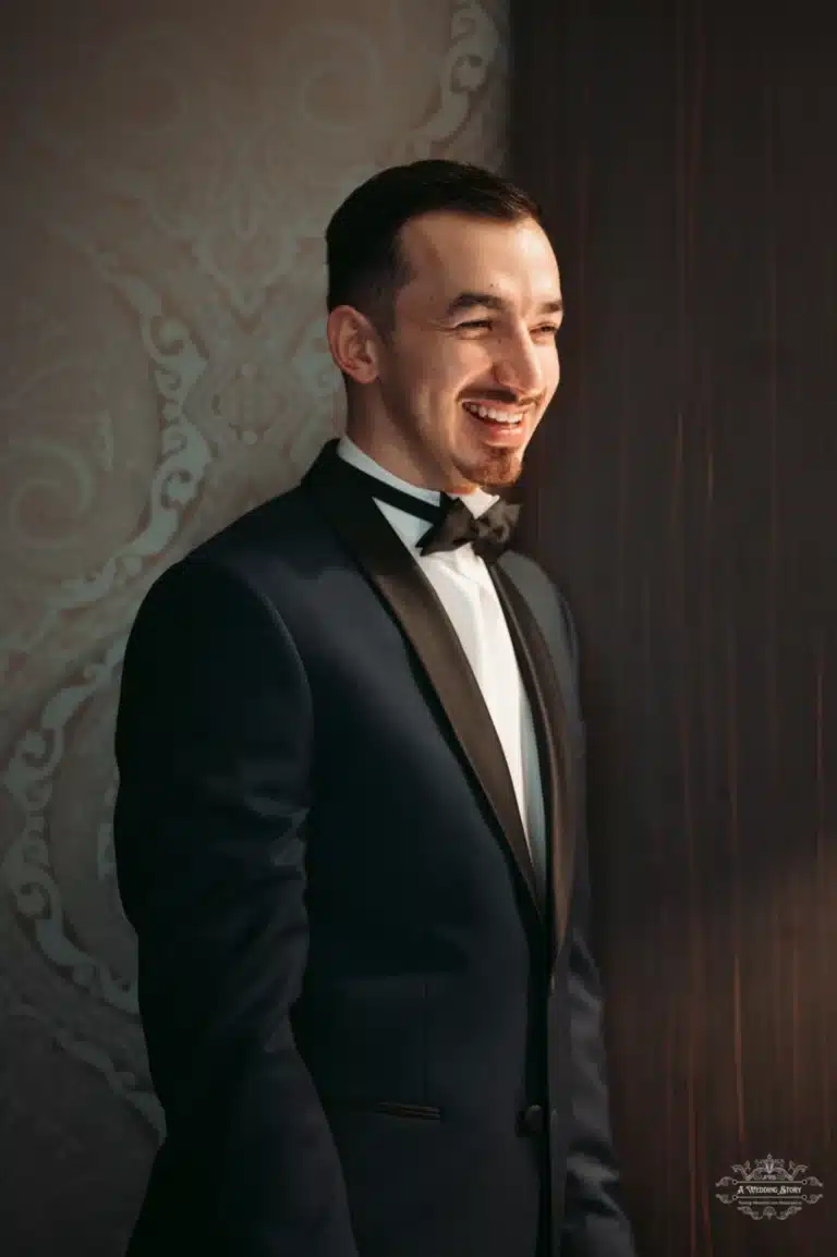 The groom, dressed in a dark tuxedo and bow tie, smiling warmly as he looks into the distance, captured in a softly lit room.