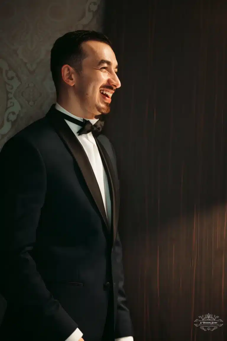 The groom in a classic tuxedo smiles warmly as he looks to the side, illuminated by soft, natural light before his wedding ceremony.