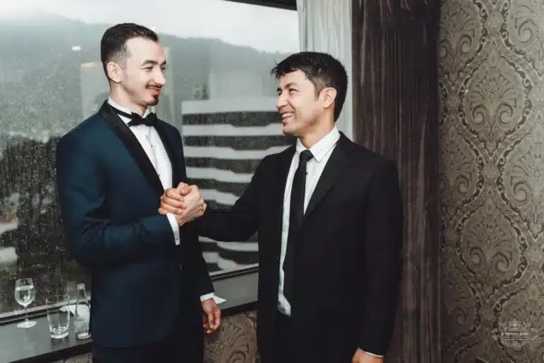 The groom in a tuxedo shares a supportive handshake with his friend before the wedding ceremony, both smiling warmly.