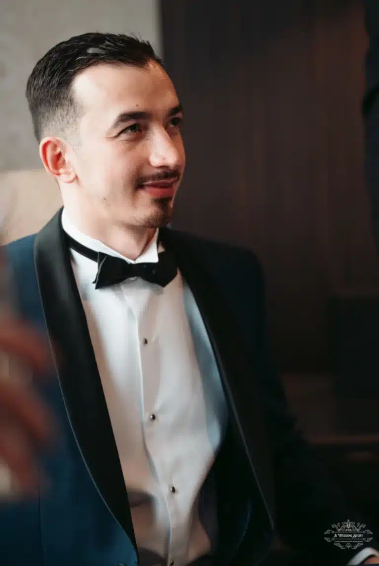 The groom, dressed in a classic tuxedo with a black bow tie, looks to the side in a thoughtful expression before his wedding ceremony.