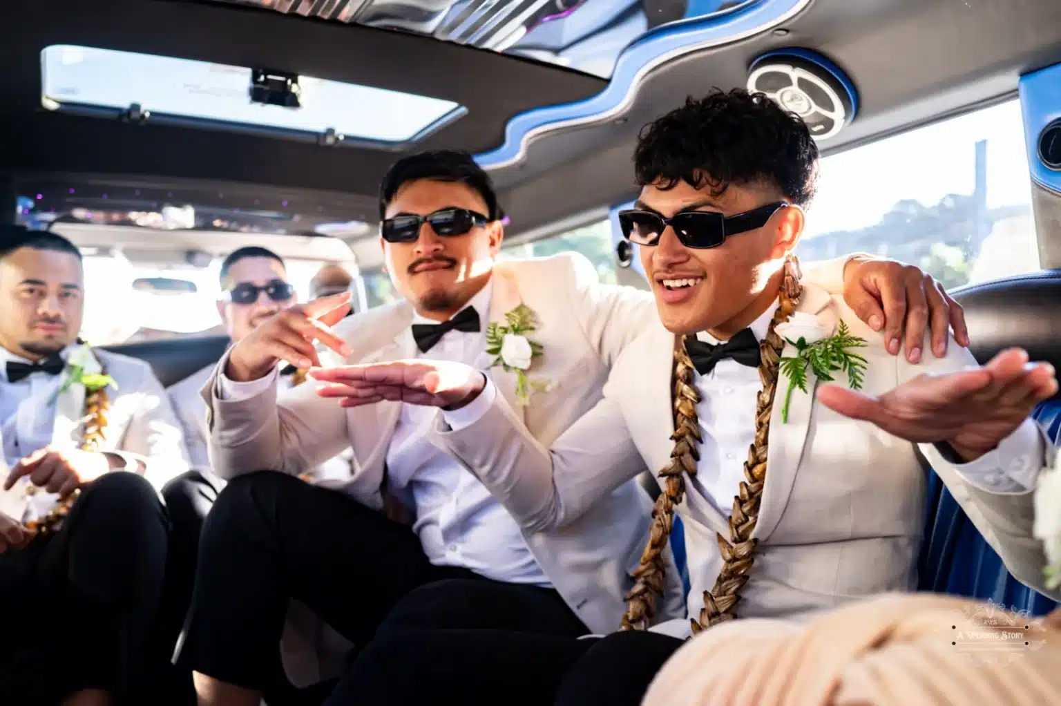 Groomsmen posing playfully inside a luxurious limousine, wearing matching white suits, bow ties, floral boutonnières, and sunglasses, with smiles and hand gestures.