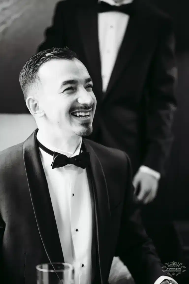 A groom smiling joyfully, dressed in a black tuxedo, with his best man standing behind him before the wedding ceremony.